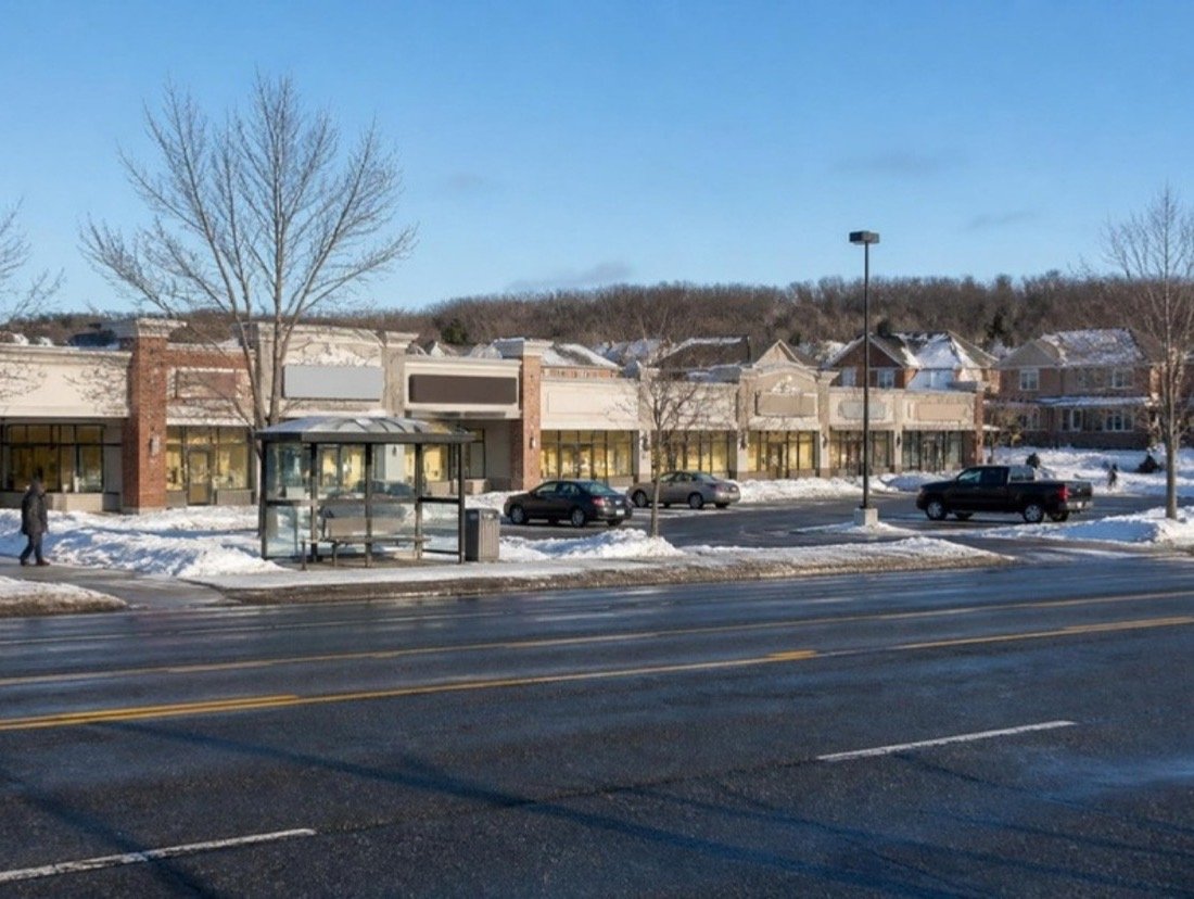 A single-storey plaza of small businesses set back from a wide arterial road on a bright winter morning, snowbanks edging the ploughed lot