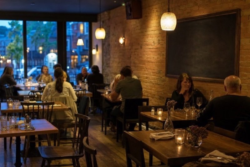 A warmly lit restaurant dining room with exposed brick walls, pendant lamps, diners at wooden tables and big windows onto a street at dusk