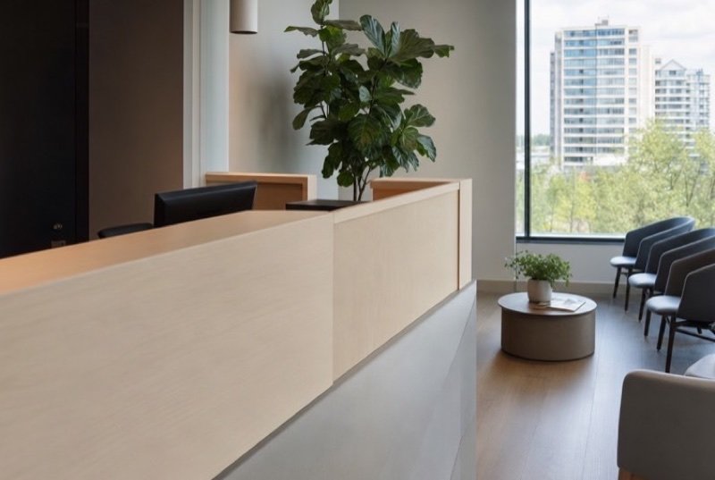 A light-filled waiting area with a wooden front desk, a tall potted plant, a row of chairs and a view of trees and mid-rise blocks