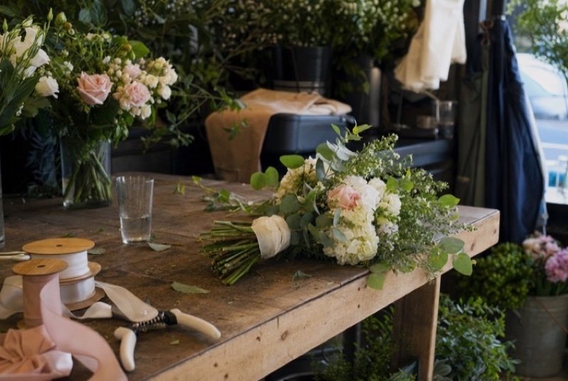 A bouquet taking shape on a wooden work table among trimmed stems, scissors and rolls of ribbon