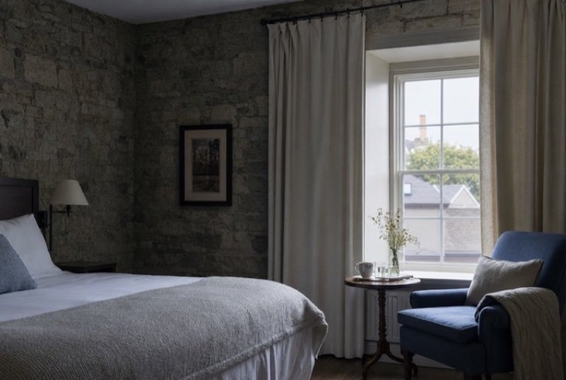 Morning sun falling across a tidy hotel bed, an armchair for reading and a bare stone wall