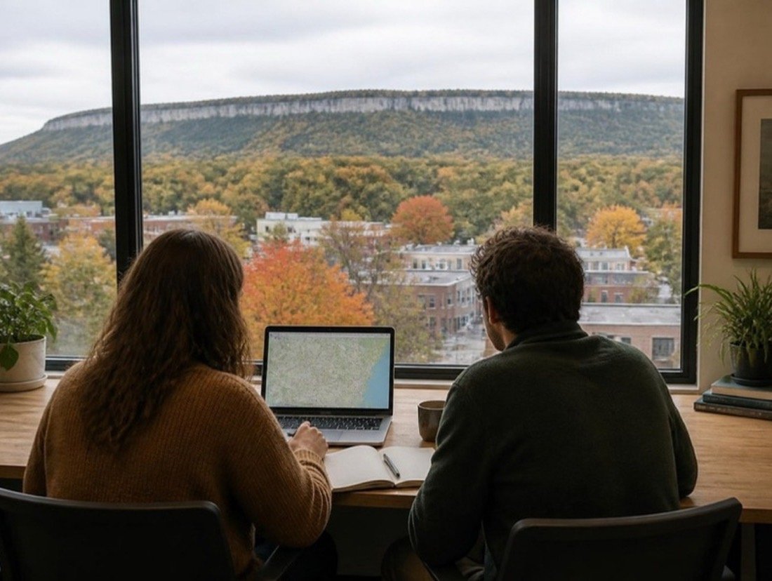 Two people seen from behind at a wooden table with a laptop and notebook, looking over rooftops and autumn maples toward a wooded ridge
